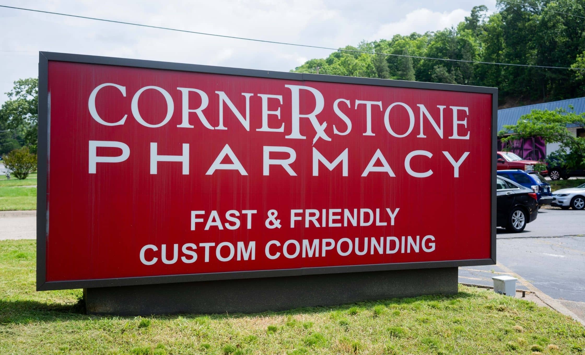 A red sign for Cornerstone Pharmacy with white text reads: Fast & Friendly Custom Compounding. The sign is outdoors near a parking lot with a few cars and greenery in the background.
